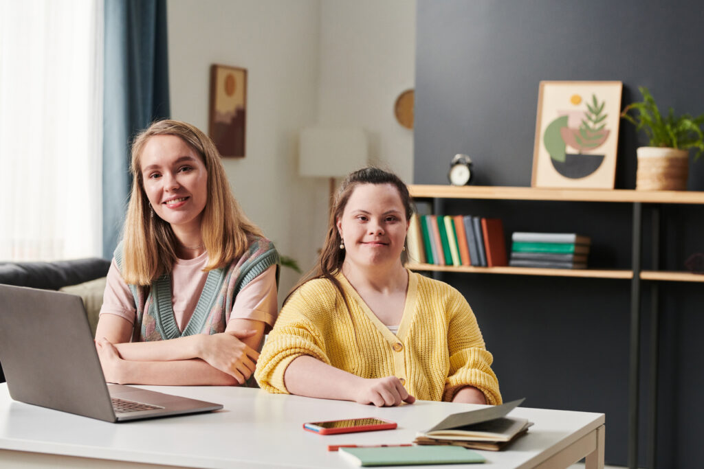 Portrait of young woman with Down syndrome and her female teacher sitting at table with laptop on it at home looking at camera.