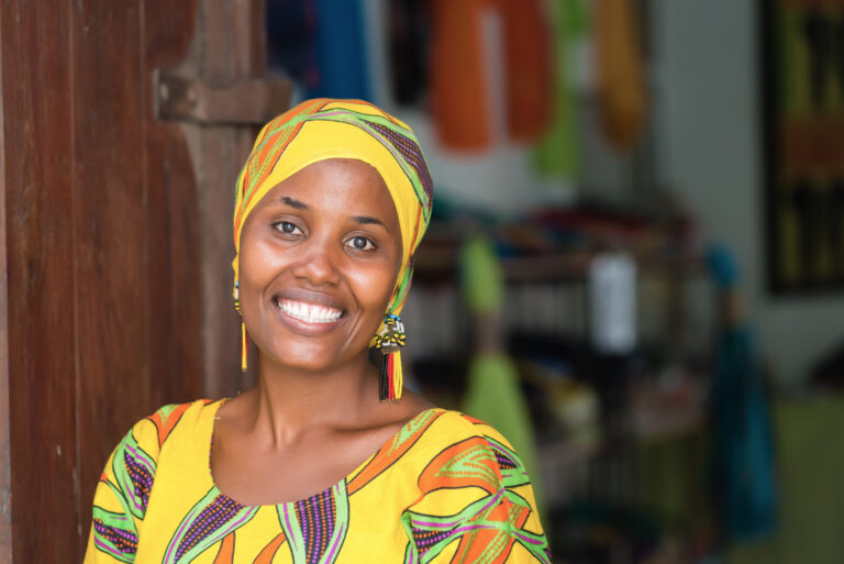 Young African woman in traditional clothes standing in entrance of her store, smiling at camera